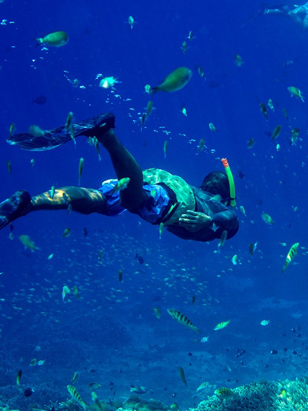 A man is scuba diving through a reef with fish around him. One of several types of excursions on t4ropical vacations brought to you by Travel Works Vacations of Carthage, MO.